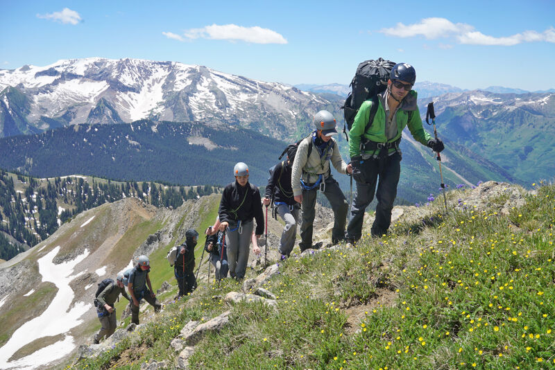 The image shows a group of hikers ascending a mountain trail. They are equipped with backpacks, hiking poles, and appropriate attire for the climb. The background features a stunning mountain range with snow-capped peaks and a clear blue sky. The hikers appear to be enjoying the scenic views and the challenge of the hike.